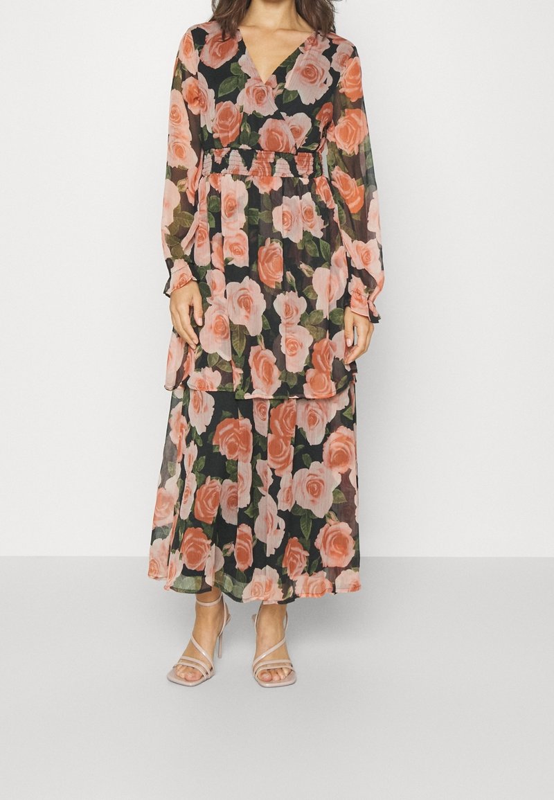 Femme portant une longue robe noire avec de grandes impressions de roses roses et pêche, des manches longues transparentes et des sandales à talons hauts beiges à brides fines.
