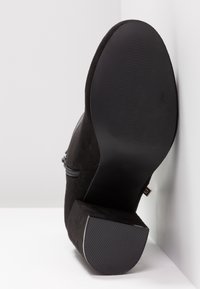 Botte cheville en daim noir avec talon bloc, semelle en caoutchouc visible et fermeture éclair latérale, positionnée semelle vers le haut contre une surface blanche.