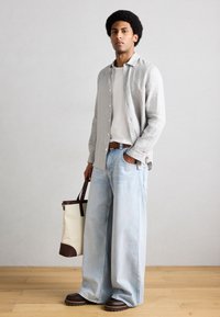 Άνδρας που στέκεται μέσα σε εσωτερικό χώρο, φοράει φαρδιά ανοιχτόγαλατζια τζιν, λευκό πουκάμισο, ανοιχτό γκρι πουκάμισο με κουμπιά, καφέ παπούτσια και κρατάει μπεζ τσάντα tote με καφέ λαβές.