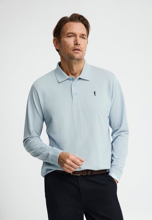 Homme portant un polo à manches longues bleu clair avec un petit logo de golfeur, un pantalon foncé et une ceinture marron, debout avec une main dans la poche.