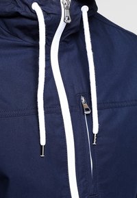 Marin blå vindjacka med vita dragsnören, framdragkedja och bröstficka. Har en slät yta och en sportig design.