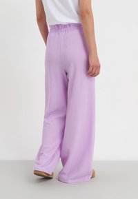 Pantalons à jambes larges en lavande claire, dotés d'un tissu texturé et d'une taille élastique. L'ourlet tombe près du sol, associés à des chaussures plates.