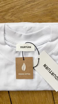 Valge orgaanilisest puuvillast T-särk kenasti kokku volditud. Siltide seas on "YOURTURN" label ja pruun silt lehelogoga, millel on kirjas "ORGANIC COTTON."