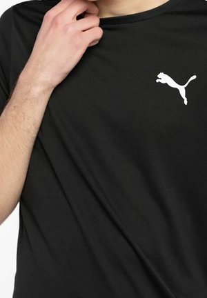 Osoba ubrana w czarną koszulkę marki Puma, trzymająca prawą ręką za kołnierz, ukazując białe logo Puma na piersi.