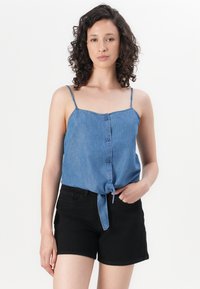 Blusa di denim azzurro chiaro con chiusura frontale a bottoni e orlo annodato, abbinata a pantaloni corti neri. Spalline sottili regolabili e vestibilità rilassata.