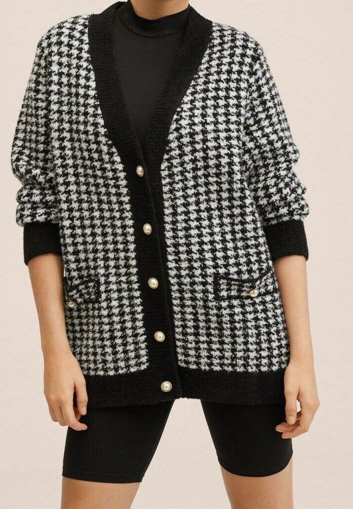 Cardigan pied-de-poule noir et blanc avec bordure noire et boutons en perle, porté sur un haut à manches longues noir et un short de cycliste noir.