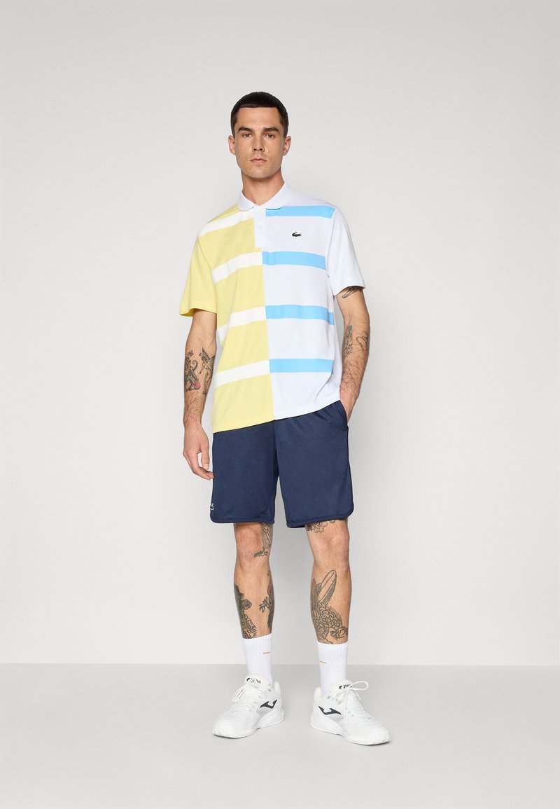 Le polo présente des rayures horizontales jaunes, blanches et bleues, en coton, avec un col classique. Il est associé à un short marine et des baskets blanches.