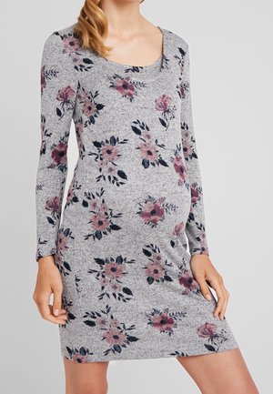 Frau trägt ein graues, langärmeliges Umstandskleid mit Blumenmuster in Pink und Dunkellila, hält das Kleid auf Oberschenkelebene.