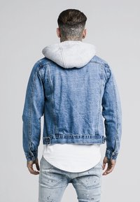 Jeansjacka med ljusblå tvätt, med en grå huva, slim fit och subtila stygn. Bärs över en lång vit skjorta.