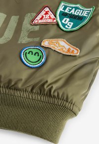 Veste bomber vert olive avec poignets côtelés, ornée de plusieurs écussons brodés colorés : un visage souriant, un triangle, un écusson et un logo.