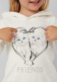 Un hoodie blanc avec une illustration de deux chatons moelleux aux yeux bleus, encadrée par un ruban gris, et le mot "AMIS" en strass.