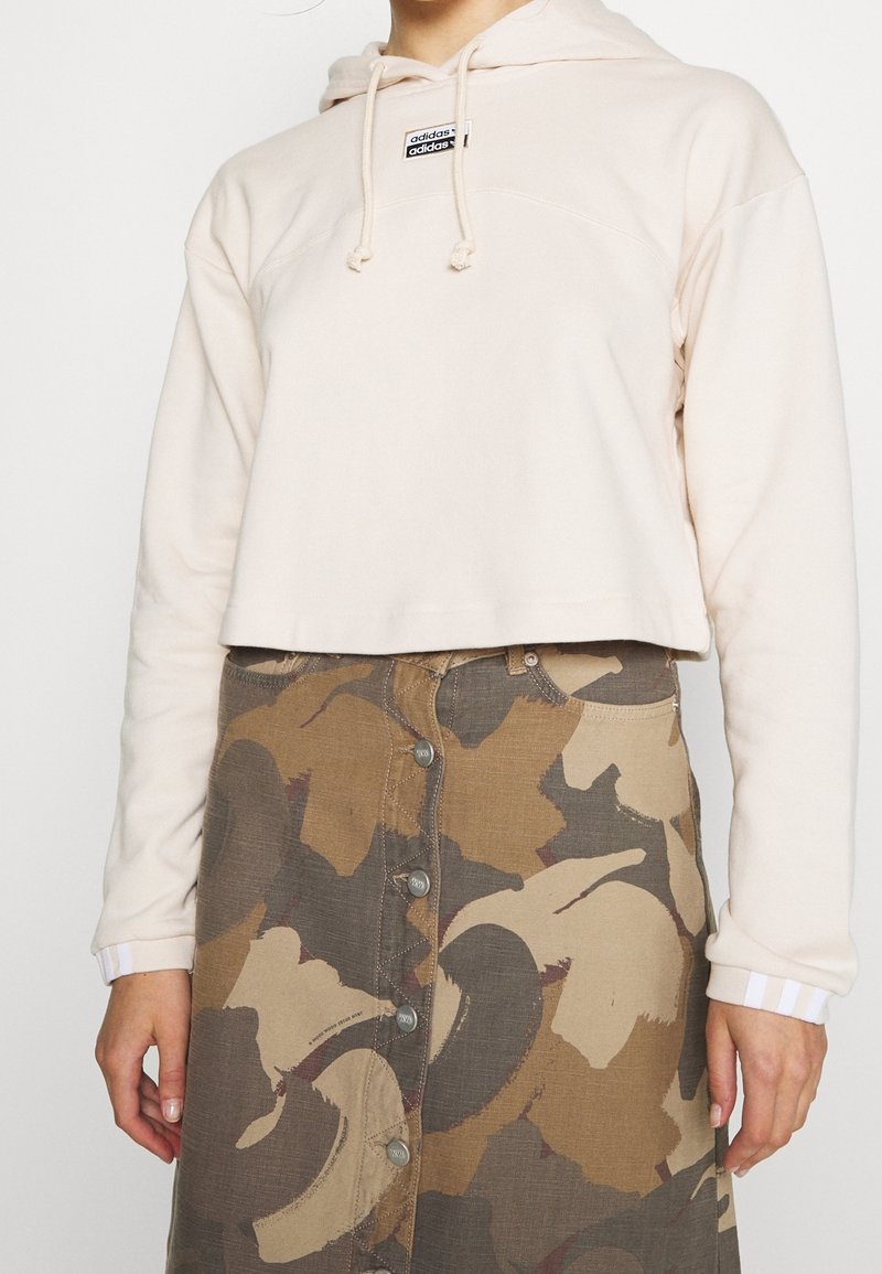 Cropped beige hoodie with drawstrings and logo, paired with a camo-pattern skirt featuring buttons and pockets in earth tones.