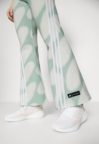 Ljusgröna utsvängda byxor med ett slätt mönster och vita sidostråk, kombinerat med vita träningsskor som har en texturerad mesh-design.