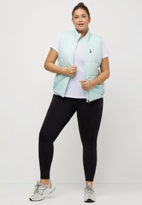 Gilet imbottito azzurro chiaro con alto colletto, chiusura a zip e tasche laterali, indossato sopra una t-shirt bianca e abbinato a leggings neri e sneakers.