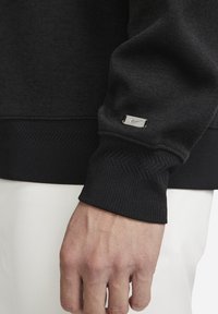 Svart sweatshirt med strukturerad tyg, ribbade ärmslut och en silverfärgad Nike-logotag på ärmen, med ett fiskbensmönster på ärmslutet.