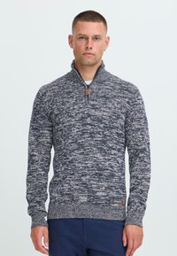 Hombre con cabello corto que lleva un suéter de punto jaspeado en blanco y negro con un cuello de cremallera de un cuarto y pantalones azul marino, de pie contra un fondo blanco.