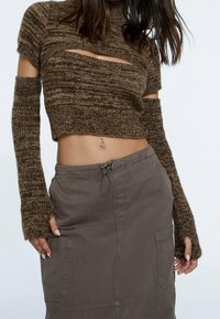 Maglione corto a maglia marrone con maniche lunghe e dettagli con taglio, abbinato a una gonna grigia con tasche e vita regolabile con cordoncino.