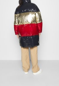 Puffjacka i marinblått, guld och röda paneler; blank yta. Kombinerad med beigefärgade byxor och vita sneakers. Har hög krage och långa ärmar.