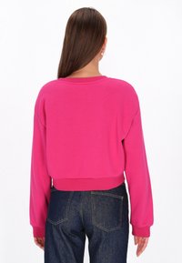 Fukszia színű, rövidített pulóver, hosszú, bő ujjakkal és bordázott mandzsettákkal. Sima textúra, kerek nyakkivágás és befejezetlen alj. Sötétkék farmerrel párosítva.