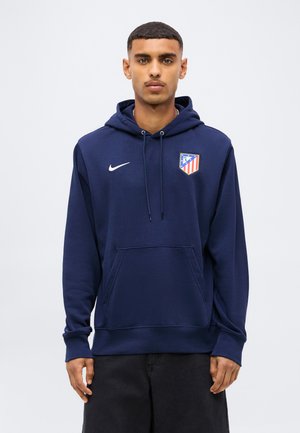 Mann trägt einen dunkelblauen Nike-Hoodie mit dem Logo des Fußballvereins Atletico Madrid und steht vor einem schlichten hellen Hintergrund.