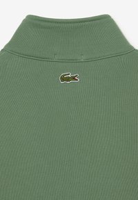 Polo verde con un tessuto a maglia texturizzato. Presenta un colletto a pieghe e un logo di coccodrillo ricamato sulla schiena vicino alla scollatura.