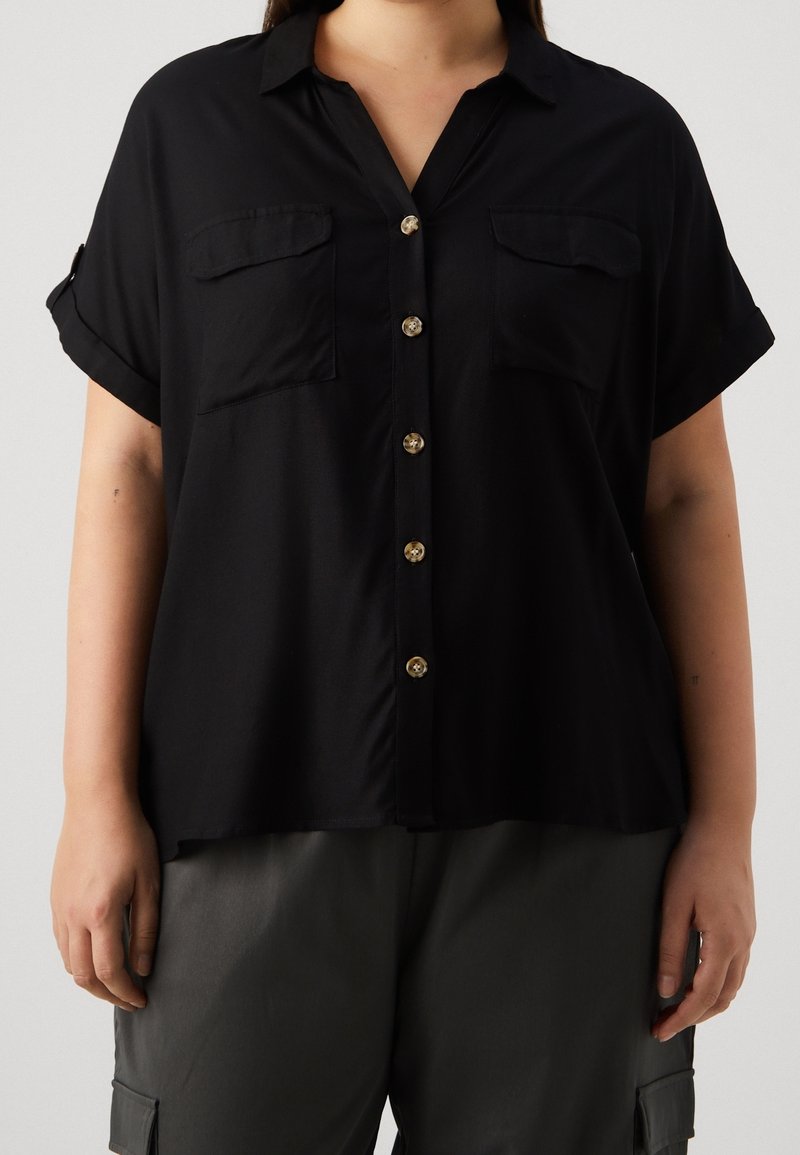 Femme portant une chemise noire à manches courtes avec des poches poitrine et un pantalon cargo gris foncé sur un fond uni.