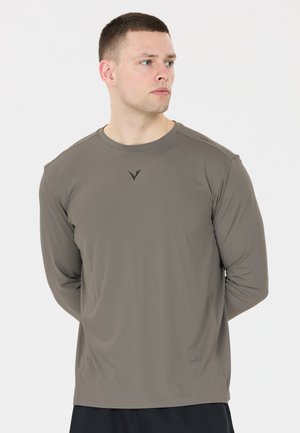 Man met kort haar die een grijs sportshirt met lange mouwen draagt, staat met handen achter de rug en kijkt opzij tegen een witte achtergrond.