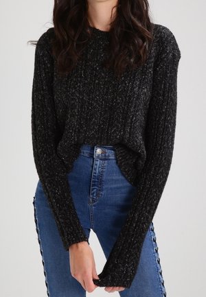 Frau trägt einen schwarzen, strukturierten Strickpullover mit langen Ärmeln und hochtaillierte blaue Jeans mit seitlichen Knopfdetails.