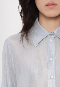 Gros plan d'une personne portant une chemise boutonnée bleu clair scintillante avec un col pointu et des cheveux foncés lâchés sur fond blanc.