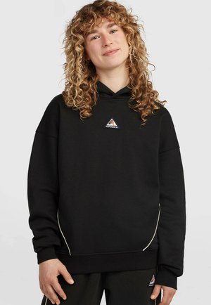 Jong persoon met krullend haar, gekleed in een zwarte O'Neill hoodie en bijpassende broek, licht glimlachend en staand tegen een effen achtergrond.