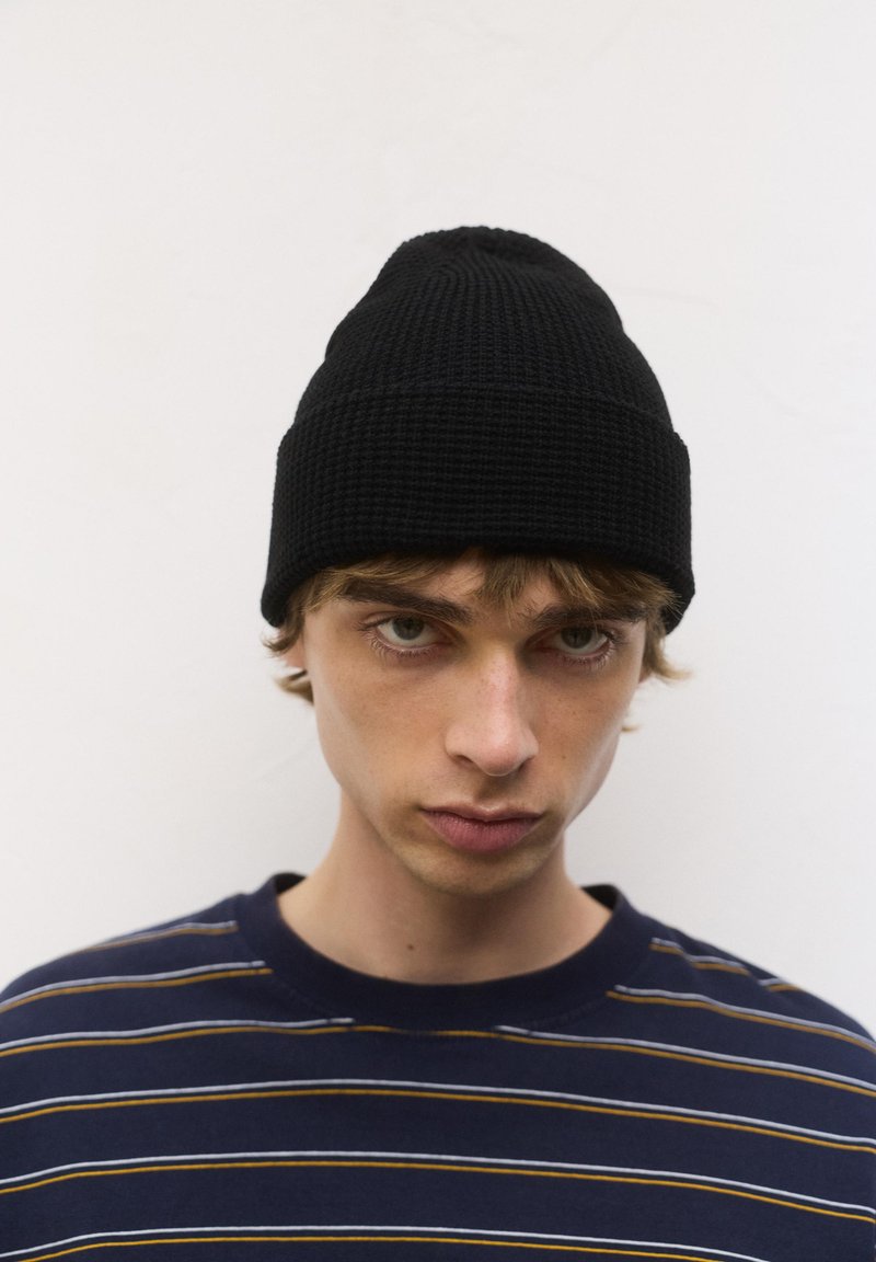 Jeune homme aux cheveux châtain clair portant un bonnet en tricot noir et un pull rayé bleu marine, faisant face à l'avant avec une expression neutre.