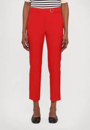 Femme portant un pantalon court rouge vif avec une ceinture assortie, une chemise rayée et des chaussures plates noires, debout devant un fond uni.