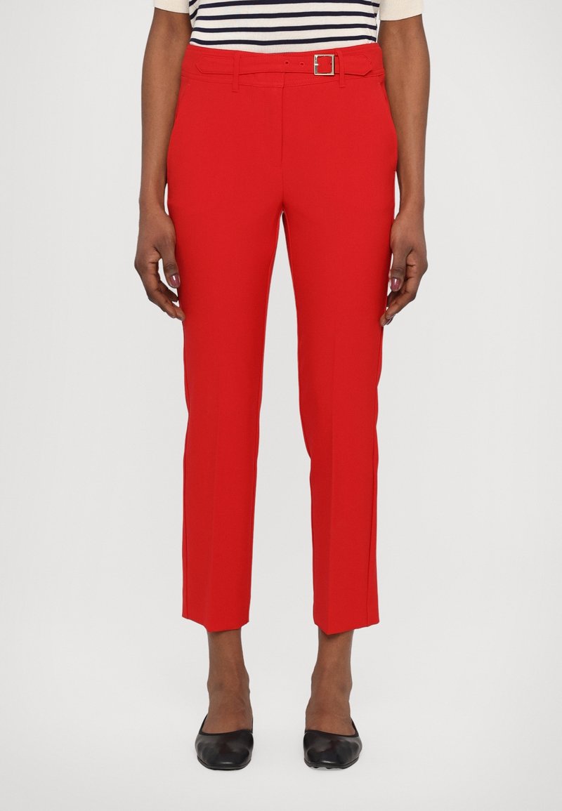 Femme portant un pantalon court rouge vif avec une ceinture assortie, une chemise rayée et des chaussures plates noires, debout devant un fond uni.