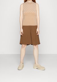 Top beige senza maniche con corpetto decorato, abbinato a pantaloni corti plissettati marroni fino al ginocchio e scarpe in camoscio di colore chiaro.