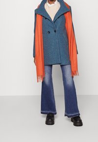 Osoba oblečená v modrém dvouřadém kabátu, s oranžovým šátkem přehozeným přes ramena, krémovém pleteném svetru, zvonových džínách a černých masivních botách.