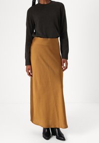 Een grijze trui gecombineerd met een mosterdkleurige, enkellange rok. De rok heeft een soepele textuur en de outfit wordt compleet gemaakt met zwarte haklaarzen.