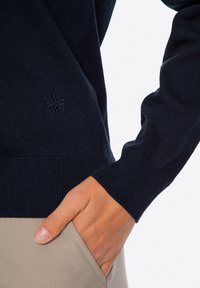 Granatowy sweter z dzianiny z prążkowanymi mankietami i dołem. Posiada subtelny detal z haftowanym logo. Ręka modelki jest umieszczona w kieszeni.
