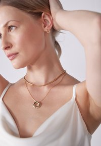 Zlatne lančiće s kružnim privjeskom složene jedan na drugi; model nosi bijelu satenastu majicu, glatku kožu i prirodan make-up, kosa je vezana.