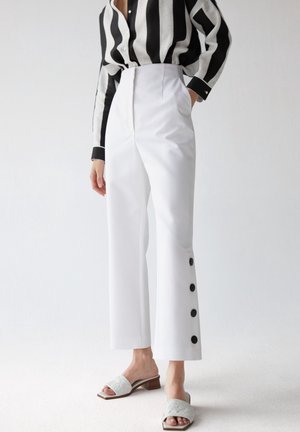 Personne portant un pantalon blanc taille haute avec des boutons noirs sur les côtés, une chemise rayée noire et blanche, et des sandales blanches à talons.