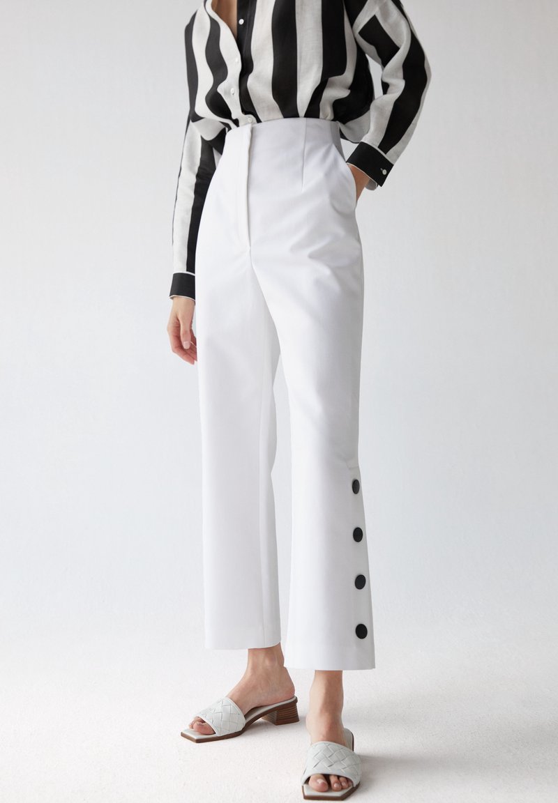 Personne portant un pantalon blanc taille haute avec des boutons noirs sur les côtés, une chemise rayée noire et blanche, et des sandales blanches à talons.