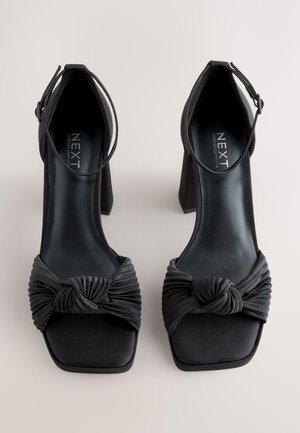 Fekete magassarkú cipő csomós selyem elülső résszel, négyzet alakú orral és vastag, strukturált sarokkal, amely állítható bokapánttal rendelkezik.