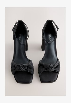 Fekete magassarkú cipő csomós selyem elülső résszel, négyzet alakú orral és vastag, strukturált sarokkal, amely állítható bokapánttal rendelkezik.