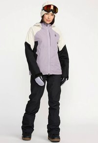 Skijakke i lavendel og creme med sorte ærmer og bukser, med lynlås foran, handsker og en strikket hue med goggles.