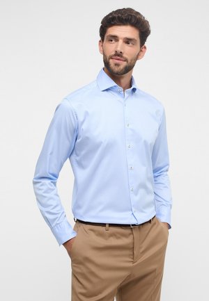 Chemise à manches longues bleu clair en tissu lisse, avec un col boutonné, des boutons sur le devant et des poignets boutonnés. Portée avec un pantalon beige.