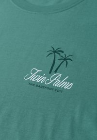 Tissu vert avec un dessin de deux palmiers et le texte « Twin Palms » et « The Barefoot Edit » sous les palmiers.