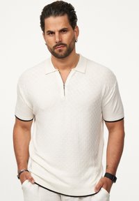 Weißes gestricktes Polohemd mit Reißverschlusskragen, kurzen Ärmeln und schwarz eingefassten Bündchen, mit einem strukturierten Muster über das gesamte Hemd.
