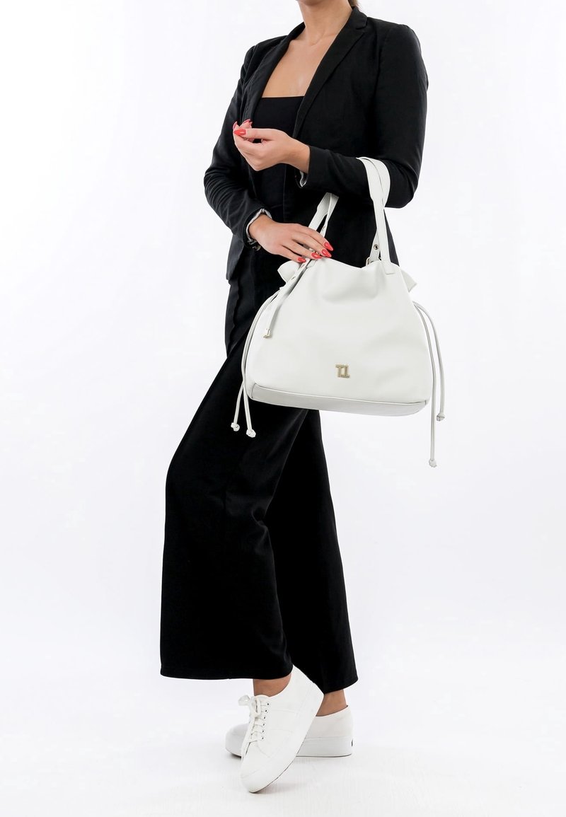Femme en tenue noire tenant un grand sac à main blanc avec double poignée, portant des baskets blanches, sur un fond uni.