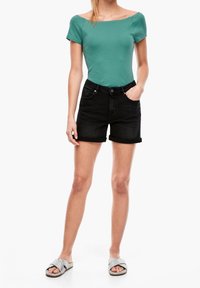 Zelený top s krátkým rukávem spárovaný s černými denimovými kraťasy s ohrnutými lemy. Model má na sobě stříbrné pantofle s texturovanými pásky.