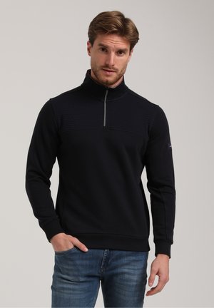 Schwarzer Zip-Up-Pullover mit strukturiertem Einsatz auf der Brust, langen Ärmeln und gerippten Bündchen. Getragen mit hellblauen Jeans.