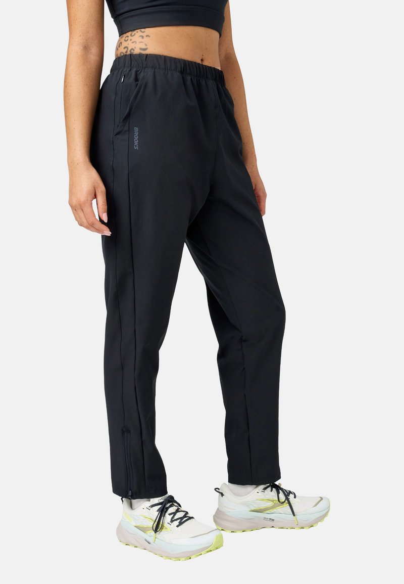 Persona vistiendo pantalones deportivos negros con cremalleras laterales, zapatillas blancas para correr y un top corto negro que revela un tatuaje parcial en la cintura.